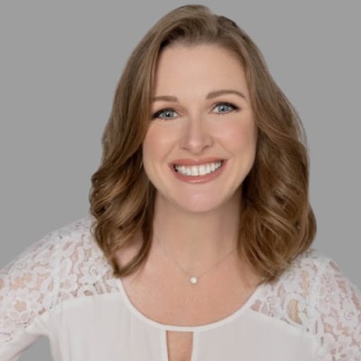 WEB_Shannon_Starline_Headshots_by_Taylor-Roman_Photography-21-removebg-preview 1 Smiling woman with shoulder-length wavy light brown hair wearing a white lace top, posing against a neutral gray background